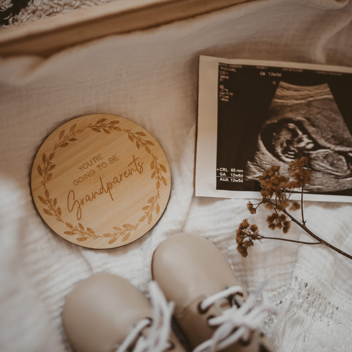 A Personalised Grandparent/Godparent Card made from bamboo is engraved with “You’re going to be grandparents” and displayed with a baby ultrasound photo, dried flowers, and baby shoes on soft cream-coloured fabric.