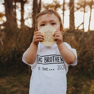 A young child outdoors holds a round card from the Lovely Lettering Pregnancy Milestones Set reading “We’re having a baby!” and wears a white shirt saying "Big brother to be... 2021," with tall trees and grass in the background.
