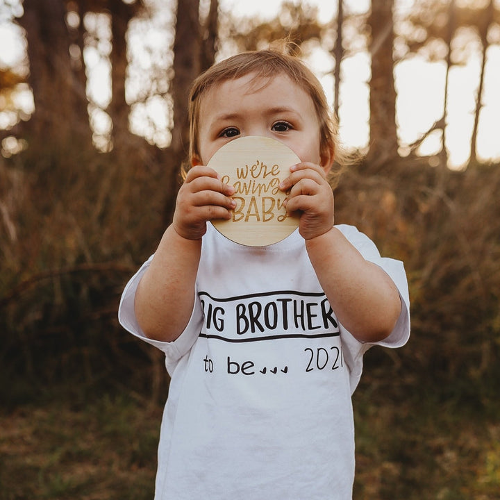 A young child outdoors holds a round card from the Lovely Lettering Pregnancy Milestones Set reading “We’re having a baby!” and wears a white shirt saying "Big brother to be... 2021," with tall trees and grass in the background.