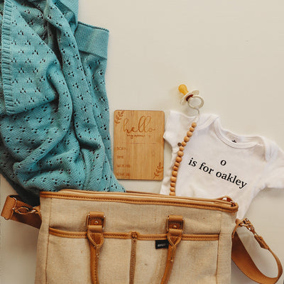 A bamboo rectangle birth announcement card saying Hello my name is. Born, Time, Weight and Length. Laying next to a baby bag and baby accessories. 