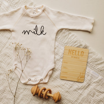 A light bamboo rectangle birth announcement card saying Hello my name is. Born, Time, Weight and Length. Laying next to a baby onesie, baby&