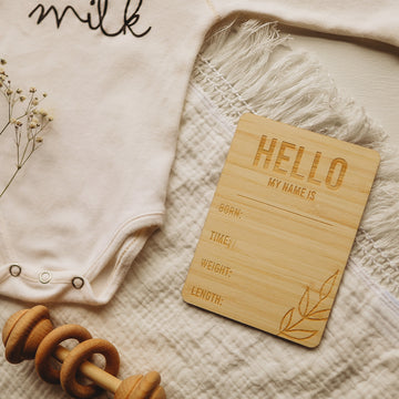 A light bamboo rectangle birth announcement card saying Hello my name is. Born, Time, Weight and Length. Laying next to a baby onesie and rattle.