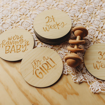 The Lovely Lettering Pregnancy Milestones Set features engraved round wooden cards with phrases like “We’re having a baby,” “It’s a girl!” and “24 weeks,” displayed on lace beside a baby rattle for the perfect milestone display.