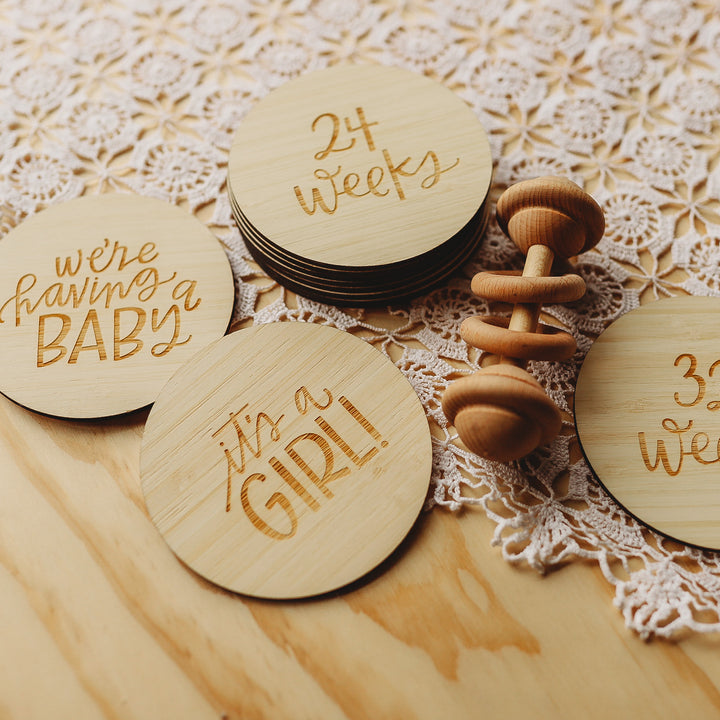 The Lovely Lettering Pregnancy Milestones Set features engraved round wooden cards with phrases like “We’re having a baby,” “It’s a girl!” and “24 weeks,” displayed on lace beside a baby rattle for the perfect milestone display.