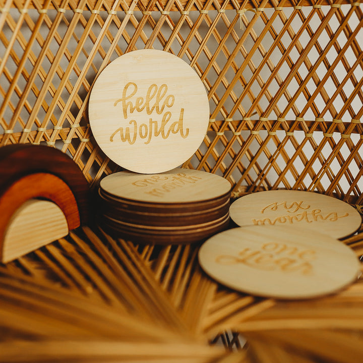 The Lovely Lettering Baby Milestone Sets include a round wooden plaque engraved with "hello world," milestone cards, and decor in warm, natural tones—perfect for documenting precious baby milestones.