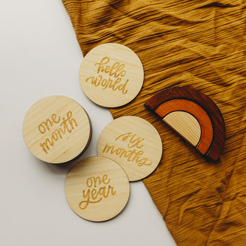 Four Lovely Lettering Baby Milestone Sets (hello world, one month, six months, one year) are displayed on mustard yellow cloth next to a wooden rainbow toy.