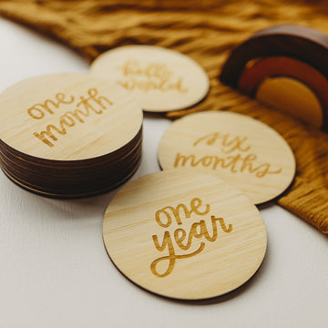 Lovely Lettering Baby Milestone Sets features round wooden cards with engraved milestones—one month, six months, and one year—arranged with a card holder and brown fabric on a white surface, perfect for capturing baby’s special moments.