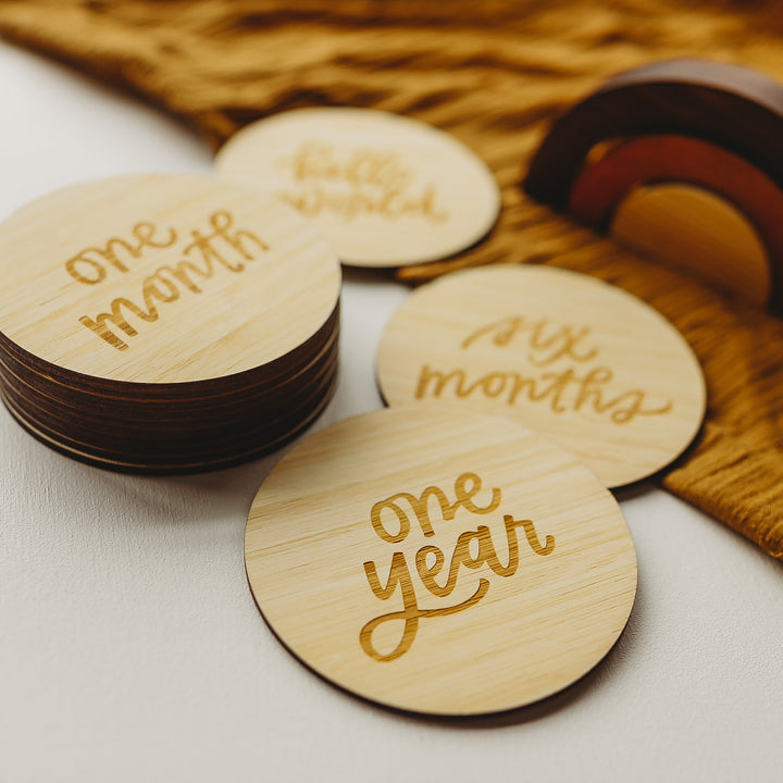 Lovely Lettering Baby Milestone Sets features round wooden cards with engraved milestones—one month, six months, and one year—arranged with a card holder and brown fabric on a white surface, perfect for capturing baby’s special moments.