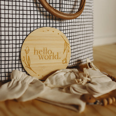 Round bamboo plaque saying Hello World with a floral border. Sitting in front of a nappy bag.