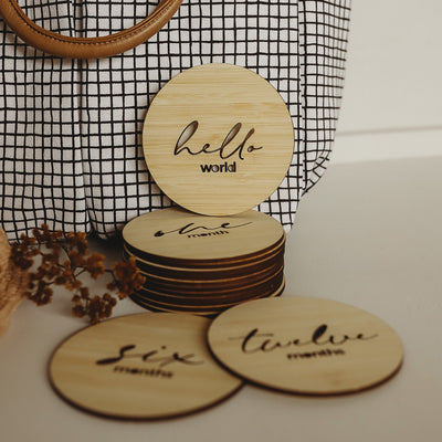 A round light bamboo set of milestone markers, sitting in front of a black and white nappy bag. Visible signs say Hello world, one month, six months and twelve months. 