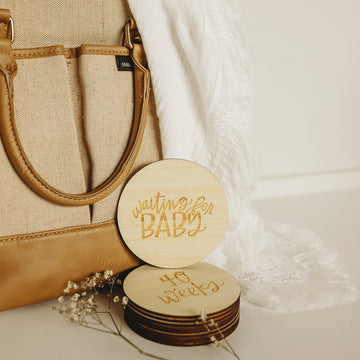 A tan and brown handbag sits next to a white fringed blanket. In front, the Lovely Lettering Pregnancy Milestones Set features stacked round wooden cards engraved with "waiting for BABY" and "40 weeks," accented by small flowers.