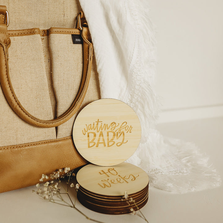 A tan and brown handbag sits next to a white fringed blanket. In front, the Lovely Lettering Pregnancy Milestones Set features stacked round wooden cards engraved with "waiting for BABY" and "40 weeks," accented by small flowers.