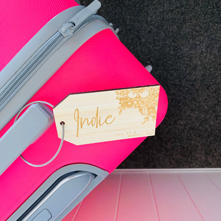 A bright pink suitcase features a Personalised Luggage Tag with a bamboo finish labelled "Indie" in cursive, attached by a silver string. The floral tag displays subtle contact details for easy identification.