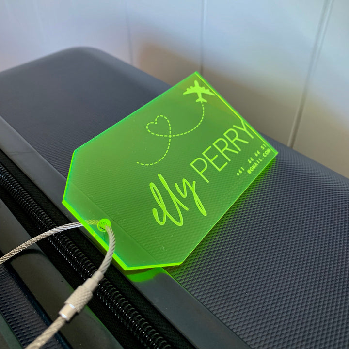 A neon green Personalised Luggage Tag labelled "My PERRY" with a heart and airplane graphic is attached to a dark textured suitcase handle by a metal cable.