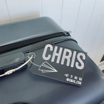 A close-up of a black suitcase featuring the Personalised Luggage Tag, which shows the name CHRIS in bold letters, a paper airplane design, and partial contact info like an email address and phone number.