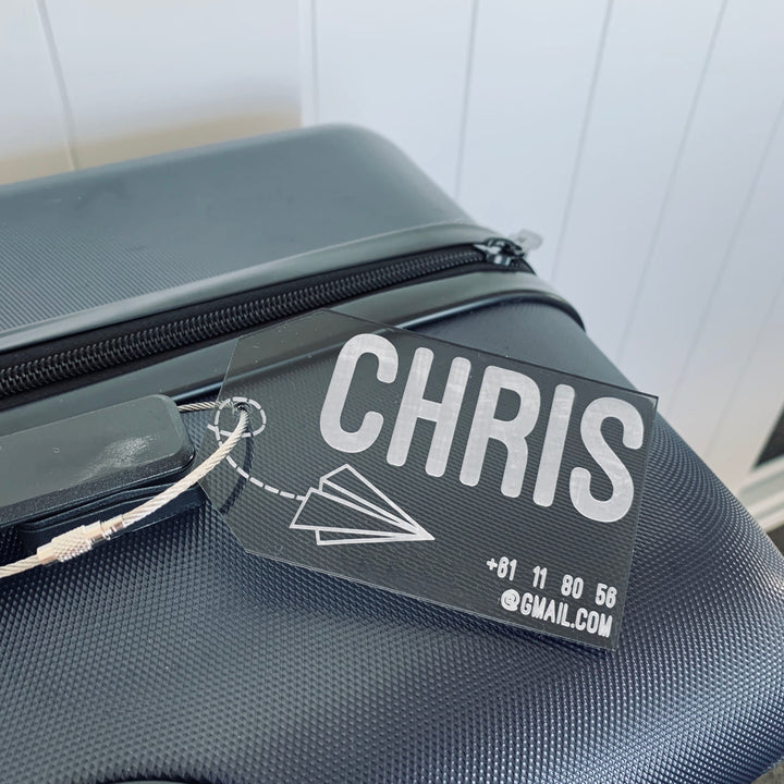 A close-up of a black suitcase featuring the Personalised Luggage Tag, which shows the name CHRIS in bold letters, a paper airplane design, and partial contact info like an email address and phone number.