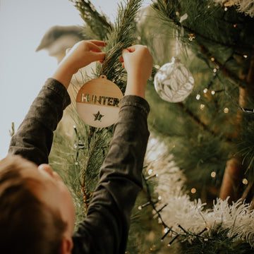 A child hangs The Original Personalised Christmas Bauble labeled HUNTER on a tree with white garland and a silver bauble—a heartwarming family tradition captured as little hands reach up to decorate.