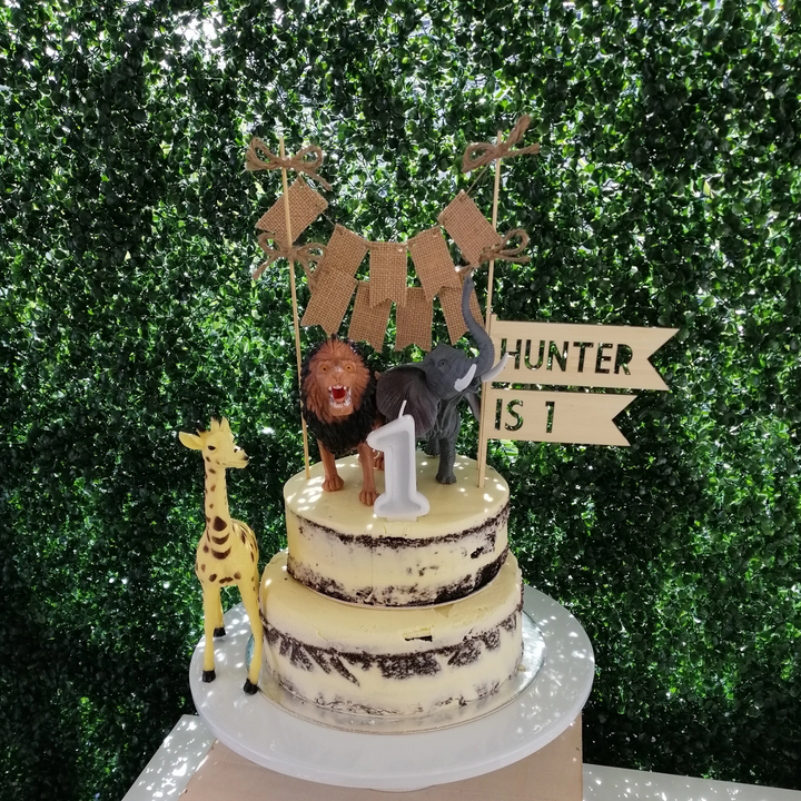 A light bamboo flag shaped cake topper saying Hunter is 1. Sitting a top a two layer cake with a giraffe, lion and elephant figurines and a 1 shaped candle. 
