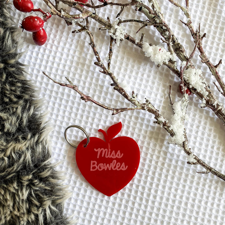 A red, apple-shaped Teacher Key Ring engraved with "Miss Bowles" sits on a white textured surface beside a snowy branch, red berries, and gray faux fur—making it a perfect personalized teacher gift.