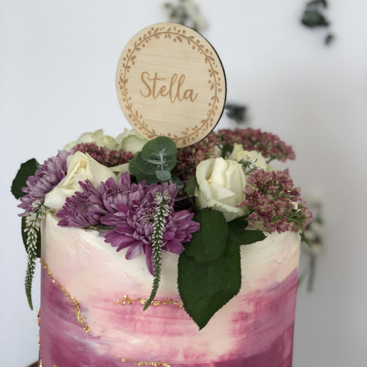 A round dark bamboo cake topper with a wreath and the name Stella engraved. Sitting on a pink and white cake with floral arrangement.