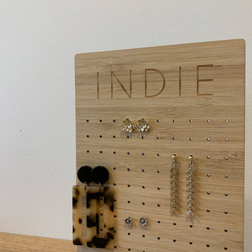 The Personalised Earring Holder, engraved with INDIE, displays floral studs, gold chain drops, and tortoiseshell and black geometric earrings against a neutral backdrop.