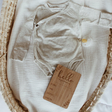 A dark bamboo rectangle birth announcement card saying Hello my name is. Born, Time, Weight and Length. Laying next to a baby onesie in a bassinet.
