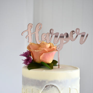 A white frosted cake is decorated with a peach rose, purple flower, green leaves, and a Mirror Personalised Cake Topper spelling "Harper" in cursive. The background is light and plain.