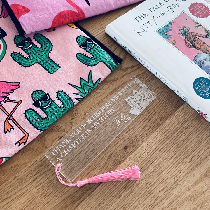 The Personalised Teacher Bookmark, featuring a clear design, pink tassel, and engraved message, sits beside cactus-patterned fabric and "The Tale of Kitty-in-Boots"—a thoughtful gift idea for teachers.