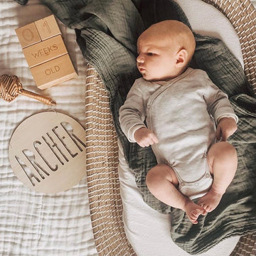 A newborn lies on a soft blanket next to a Personalised Natural Names Plaque featuring "ARCHER" and block set showing 01 weeks old, with a rattle nearby, creating a warm and cozy scene.