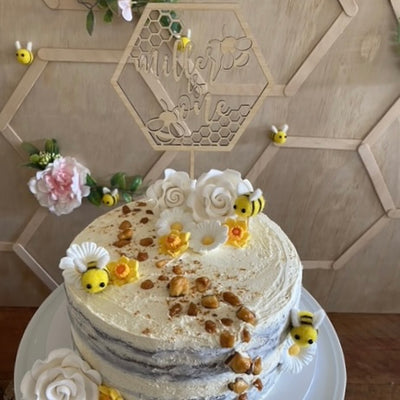 A light bamboo, hexagon shaped cake topper, with a honeycomb design, bees and Miller is one laser cut inside it. Sitting on a white cake with flowers and bumblebees. 