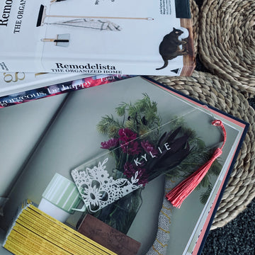 A clear bookmark etched with white florals saying Kylie, with a pink tassel, sitting a top a book of plants. 