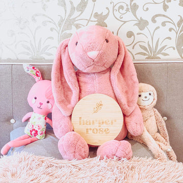 A large pink stuffed bunny holds a Personalised La Petite Stem Plaque reading "harper rose" on a couch, flanked by a pink floral bunny and beige sloth plush toy, with patterned wallpaper in the background.