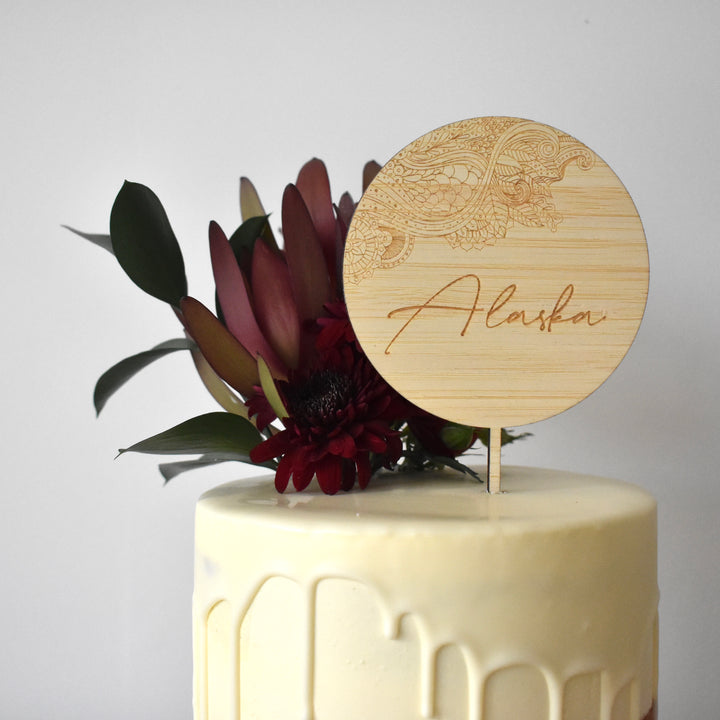 A round bamboo topper with floral design engraved and the name Alaska. Sitting atop a white drip cake with floral arrangement. 