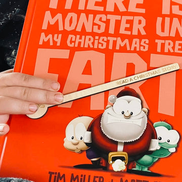 A child’s hand holds a bookmark on “There Is a Monster Under My Christmas Tree Who Farts,” with cartoon characters including Santa—a perfect companion to your Activity Advent Calendar Set for festive family fun.