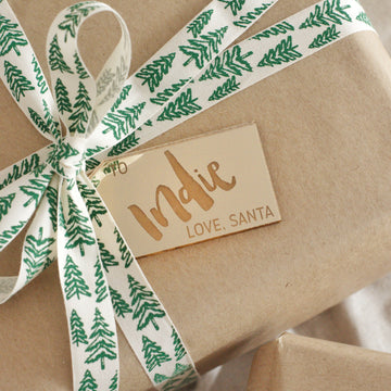A brown paper-wrapped gift with a white ribbon featuring green pine trees is topped with a gold mirrored acrylic Personalised Gift Tag reading "Indie LOVE, SANTA".