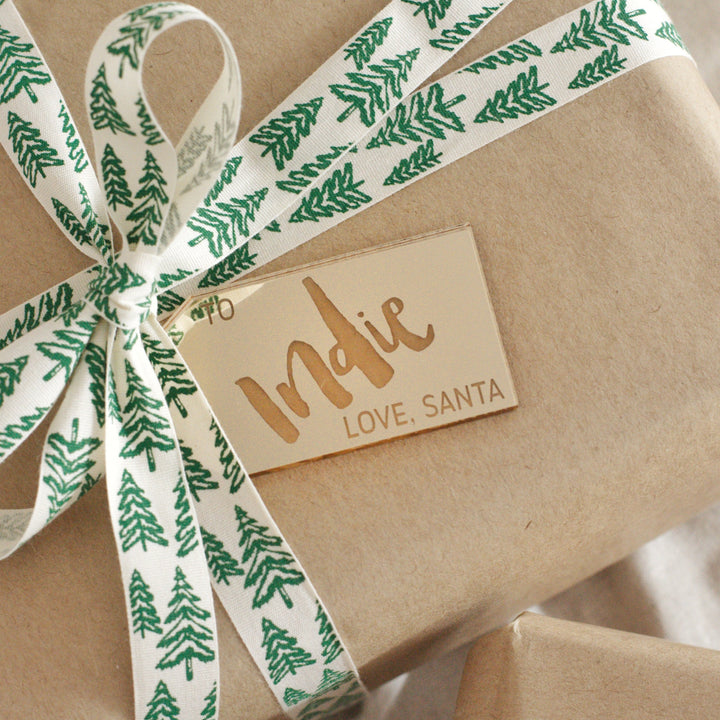 A brown paper-wrapped gift with a white ribbon featuring green pine trees is topped with a gold mirrored acrylic Personalised Gift Tag reading "Indie LOVE, SANTA".