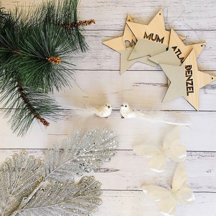 A flat lay of Christmas decorations on a white wooden surface featuring pine branches, Personalised Christmas Stars, two white decorative birds, a silver fern, bamboo ornaments, and two white butterflies.