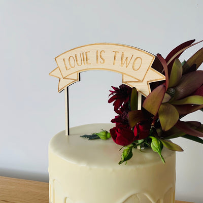 A bamboo banner cake topper saying Louie is two, sitting atop a white drip cake with floral arrangement.