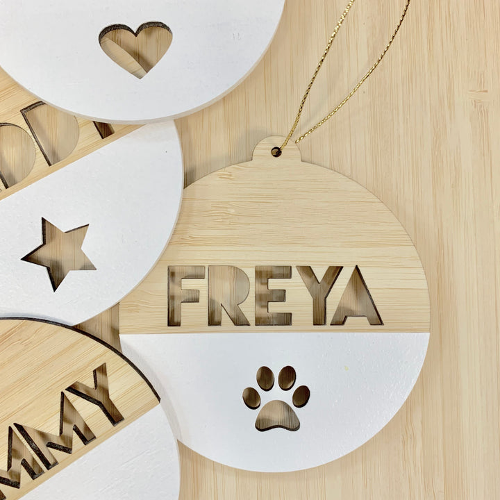 A Personalised Pet Christmas Bauble with FREYA and a paw print cutout, partly painted white, hangs on a gold string. Other custom pet ornaments with unique shapes and names are nearby on a light wood surface.