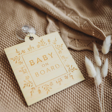 A light bamboo sign saying Baby on Board, with a border of engraved laurels and florals. Sitting atop a brown shawl and white bunny tail florals.
