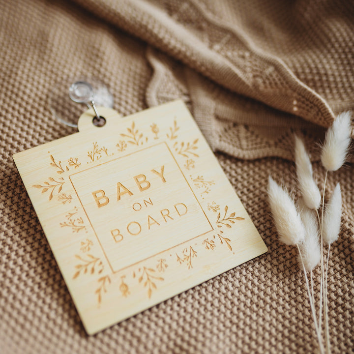 A light bamboo sign saying Baby on Board, with a border of engraved laurels and florals. Sitting atop a brown shawl and white bunny tail florals.