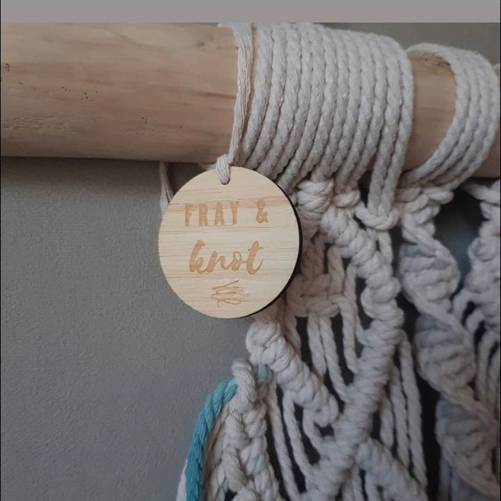 A small round light bamboo tag with Fray and Knot logo engraved, attached to a macrame piece. 
