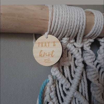 A small round light bamboo tag with Fray and Knot logo engraved, attached to a macrame piece. 