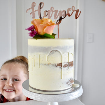 A smiling child poses next to a white-frosted, semi-naked cake with a white chocolate drip, fresh flowers, and a Mirror Personalised Cake Topper reading “Harper,” displayed on a white cake stand.