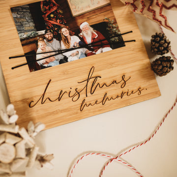 The Christmas Photo Holder, made in New Zealand, displays a family with Santa Claus. Festive decorations and red-striped twine accent the wooden plaque labeled "Christmas memories," adding holiday charm.