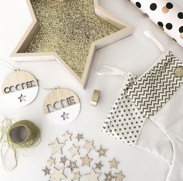 Flat lay of festive craft supplies: a star tray of gold glitter, wooden stars, two The Original Personalised Christmas Baubles, gold twine, patterned fabric, gold washi tape, and polka dot wrapping paper—ideal for a family Christmas tradition.