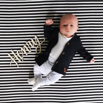 A baby lies on a black and white striped blanket, wearing a black cardigan and light pants. Beside the baby is the Lovely Lettering Handlettered Wall Name sign, elegantly spelling “Henry” in custom cursive.