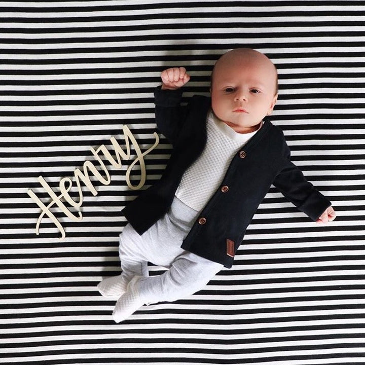 A baby lies on a black and white striped blanket, wearing a black cardigan and light pants. Beside the baby is the Lovely Lettering Handlettered Wall Name sign, elegantly spelling “Henry” in custom cursive.