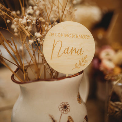 A round light bamboo planter saying In Loving Memory Nana, with a laurel engraved. Sitting with a dried floral arrangement. 