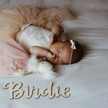 A sleeping baby in a light outfit and headband lies peacefully on a bed next to a plush toy and the Lovely Lettering Handlettered Wall Name sign that reads Birdie, with one hand near their mouth.